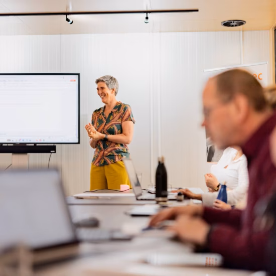 Der Trainer hält während eines Business-Workshops einen Vortrag, während die Teilnehmer mit ihren Laptops an einem Konferenztisch arbeiten.