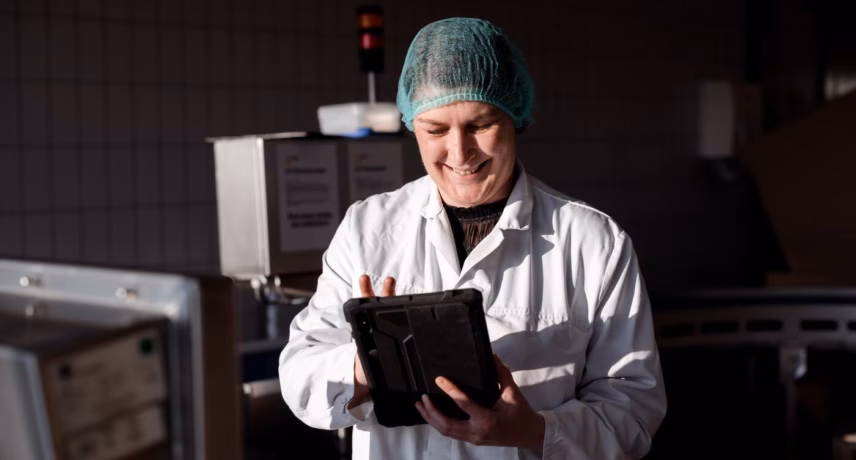 An employee in a production environment checks data on a tablet next to industrial equipment and wears protective clothing.