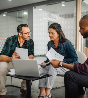 Three colleagues meeting in a modern office setting with a laptop and documents.