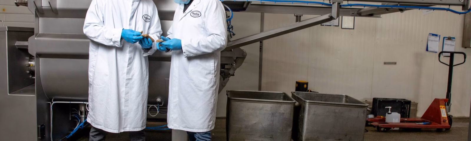 Twee mannen in witte steriele labjassen en met haarnetjes overleggen in een fabriekshal, terwijl ze productie- of kwaliteitsprocessen bespreken.