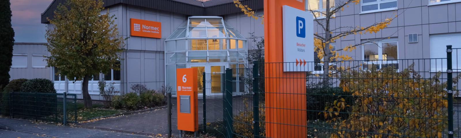Außenansicht eines modernen Forschungs- und Testzentrums mit markenspezifischer Beschilderung, einem durch ein Tor gesicherten Zugang und einem für Besucher reservierten Parkplatz, was ein professionelles Umfeld widerspiegelt, das sich wissenschaftlichen Dienstleistungen und der Qualitätssicherung widmet.