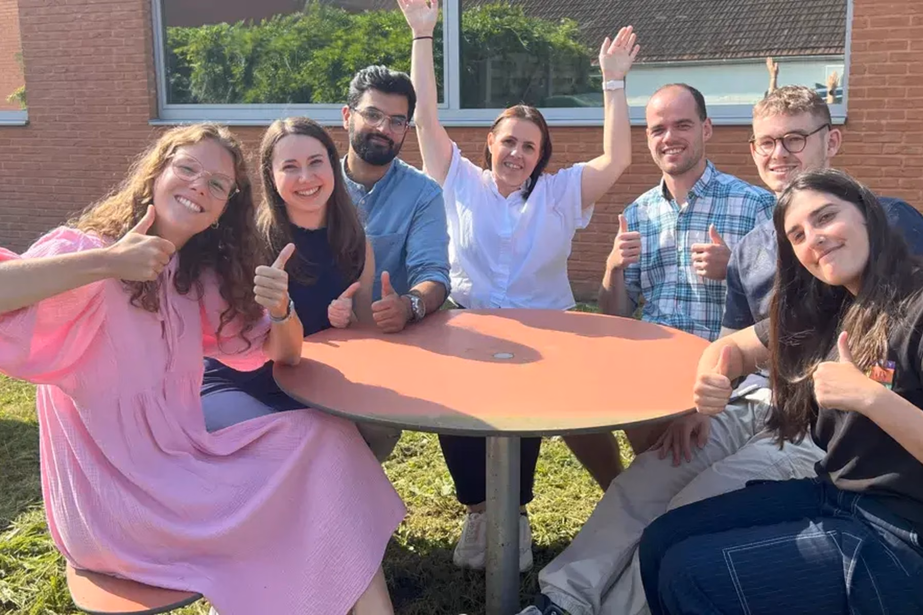 Groep collega's zit samen aan een ronde tafel op een buitenterrein bij een bedrijfspand. De afbeelding toont samenwerking, teamgevoel, kennisdeling en betrokkenheid binnen een professionele werkomgeving.