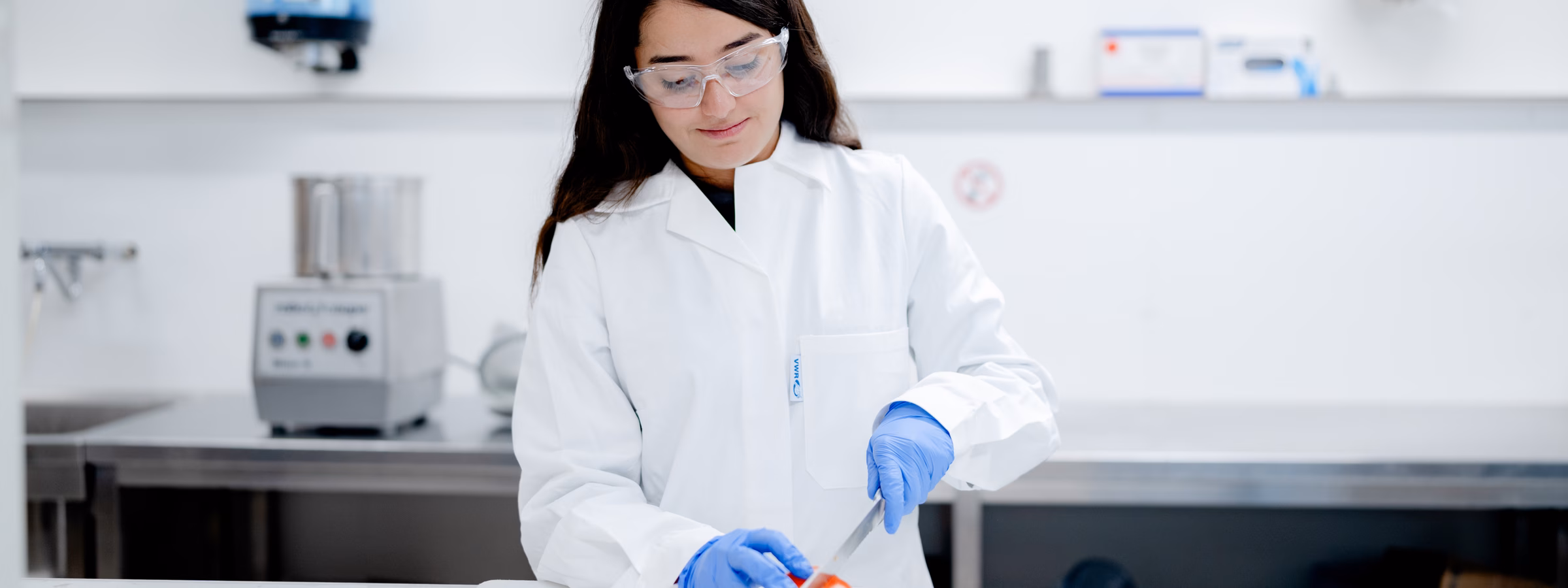 Laboratoriummedewerker onderzoekt een voedingsproduct op een werkbank in een laboratoriumomgeving. De afbeelding toont testen, kwaliteitscontrole en analyse ter ondersteuning van productveiligheid en compliance.