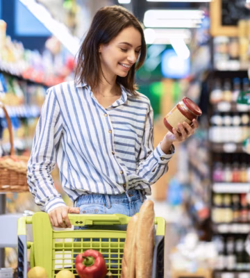 Een lachende vrouw loopt door de gangpaden van een supermarkt met een winkelwagen en bekijkt een glazen pot saus, terwijl ze gezonde producten kiest tussen de schappen.
