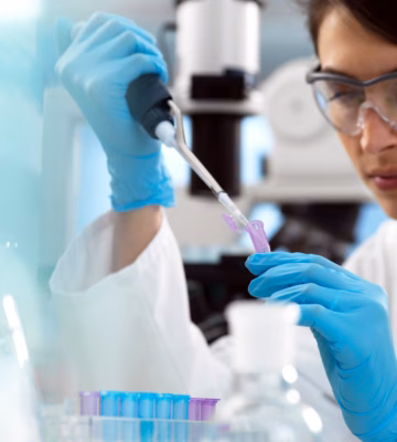 Een vrouwelijke onderzoeker met veiligheidsbril, blauwe handschoenen en witte labjas werkt geconcentreerd in het lab en pipetteert vloeistof in een reageerbuis.