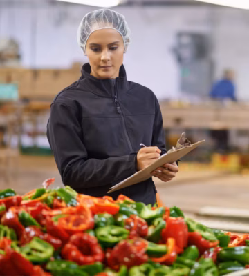 A woman wearing a hairnet is performing quality control on red and green peppers in a large bin at a vegetable processing plant, writing notes on a clipboard.