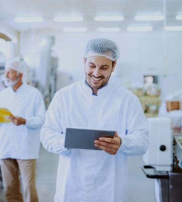 A supervisor in a food factory is wearing a white coat and hairnet, smiling while looking at his iPad and taking notes.
