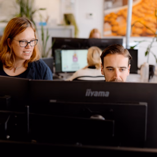 Zwei Mitarbeiter arbeiten gemeinsam vor mehreren Computerbildschirmen in einer modernen Büroumgebung.