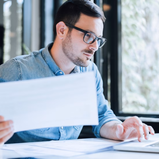 A man is typing and looking at his laptop while holding a paper in his hand, working at his desk.