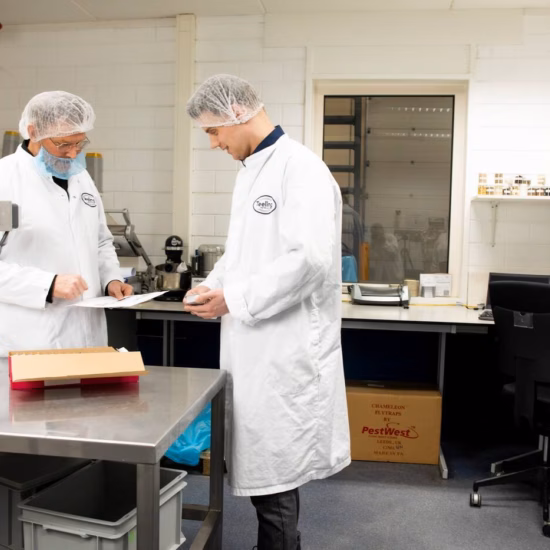 Zwei Laboranten in weißen Laborkitteln und Haarnetzen überprüfen Proben und besprechen Untersuchungsergebnisse in einem Labor.