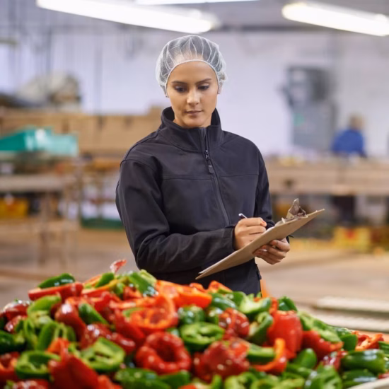 A woman wearing a hairnet is performing quality control on red and green peppers in a large bin at a vegetable processing plant, writing notes on a clipboard.