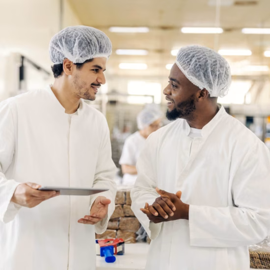 Two men are walking and smiling while conversing in a food factory, wearing sterile white coats and hairnets, with one man holding an iPad.