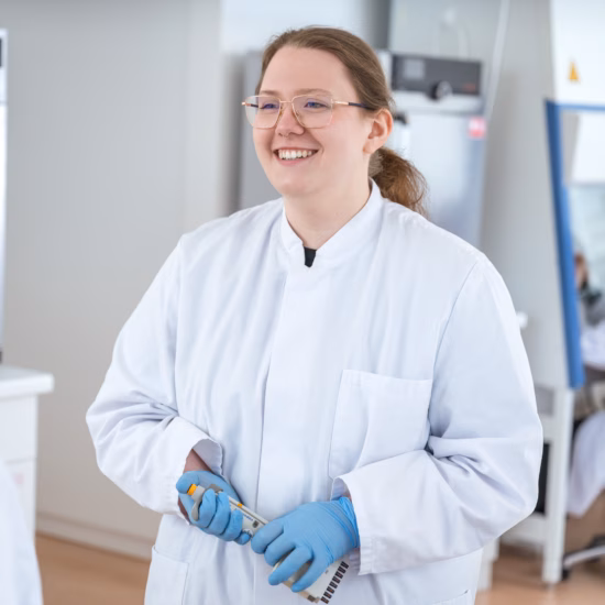 Een laboratoriumspecialist glimlacht naar een collega in een laboratorium, houdt testapparatuur vast en draagt een labjas met blauwe steriele laboratoriumhandschoenen.