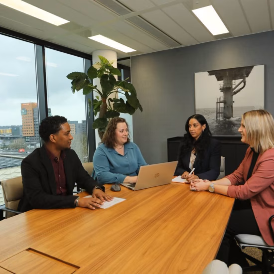 A group of four people are having a meeting in an office, chatting while one of them is writing on a laptop.