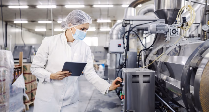 Kwaliteitsmedewerker controleert industriële productielijn in een voedselverwerkingsfabriek met tablet en hygiënische beschermingsmiddelen.