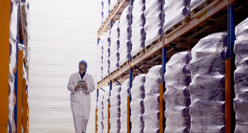 A person wearing protective work clothing walks down a warehouse aisle lined with goods stored on high racks.