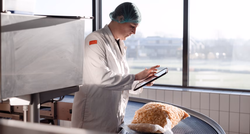 Medewerker in beschermende werkkleding controleert een broodproduct met een tablet naast een transportband in een productieomgeving.