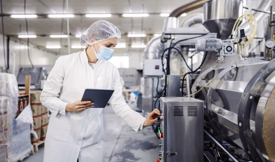 Ein Qualitätsmitarbeiter überprüft mit einem Tablet und hygienischer Schutzausrüstung eine industrielle Produktionslinie in einem Lebensmittelverarbeitungsbetrieb.
