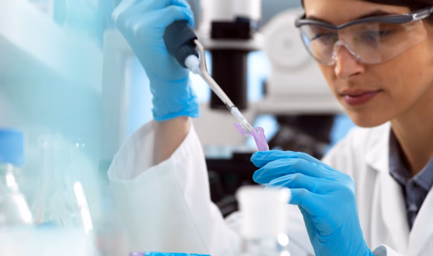Een vrouwelijke onderzoeker met veiligheidsbril, blauwe handschoenen en witte labjas werkt geconcentreerd in het lab en pipetteert vloeistof in een reageerbuis.