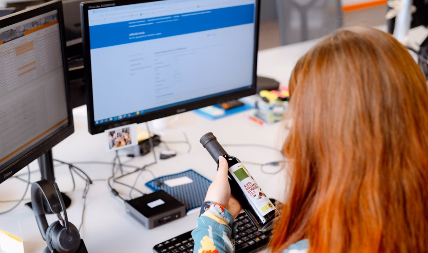 An employee uses a handheld scanner at a workstation with multiple monitors. The image illustrates digital recording, data processing, and process support within quality, inspection, or compliance processes.
