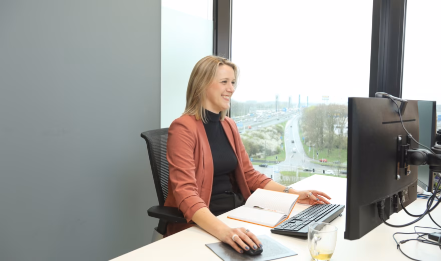 Vrouw die lacht terwijl ze op haar computer werkt, met een notitieboekje voor zich en een kopje thee.