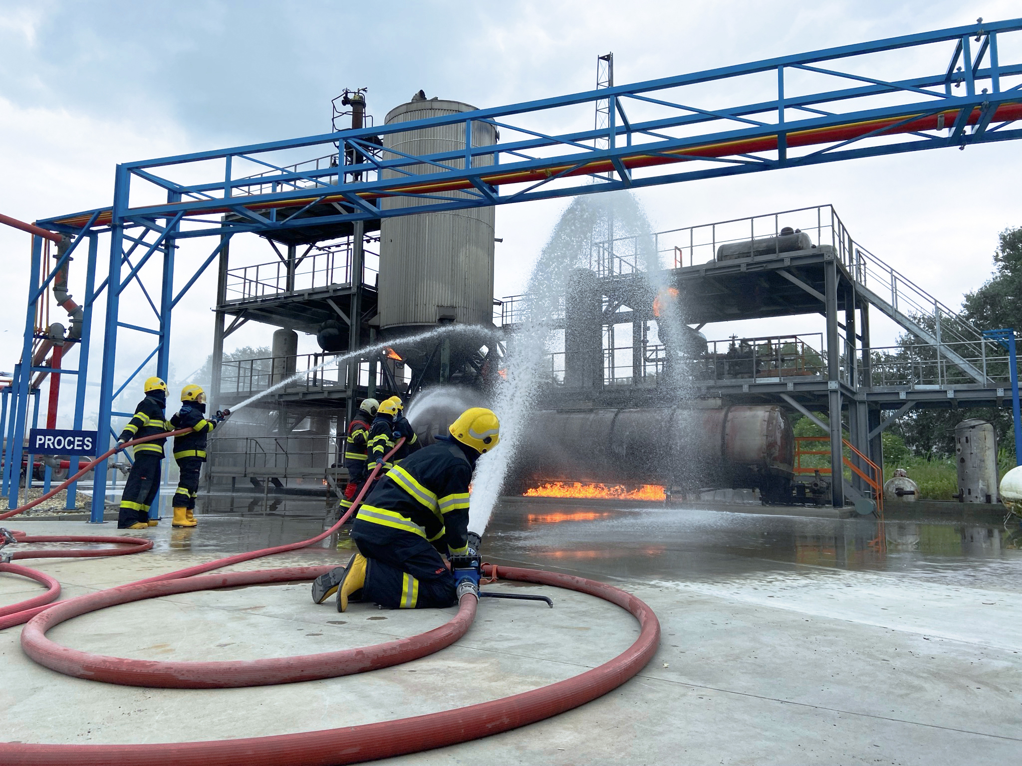 Firefighters are conducting a fire drill at an industrial facility with visible flames and water used for fire suppression.
