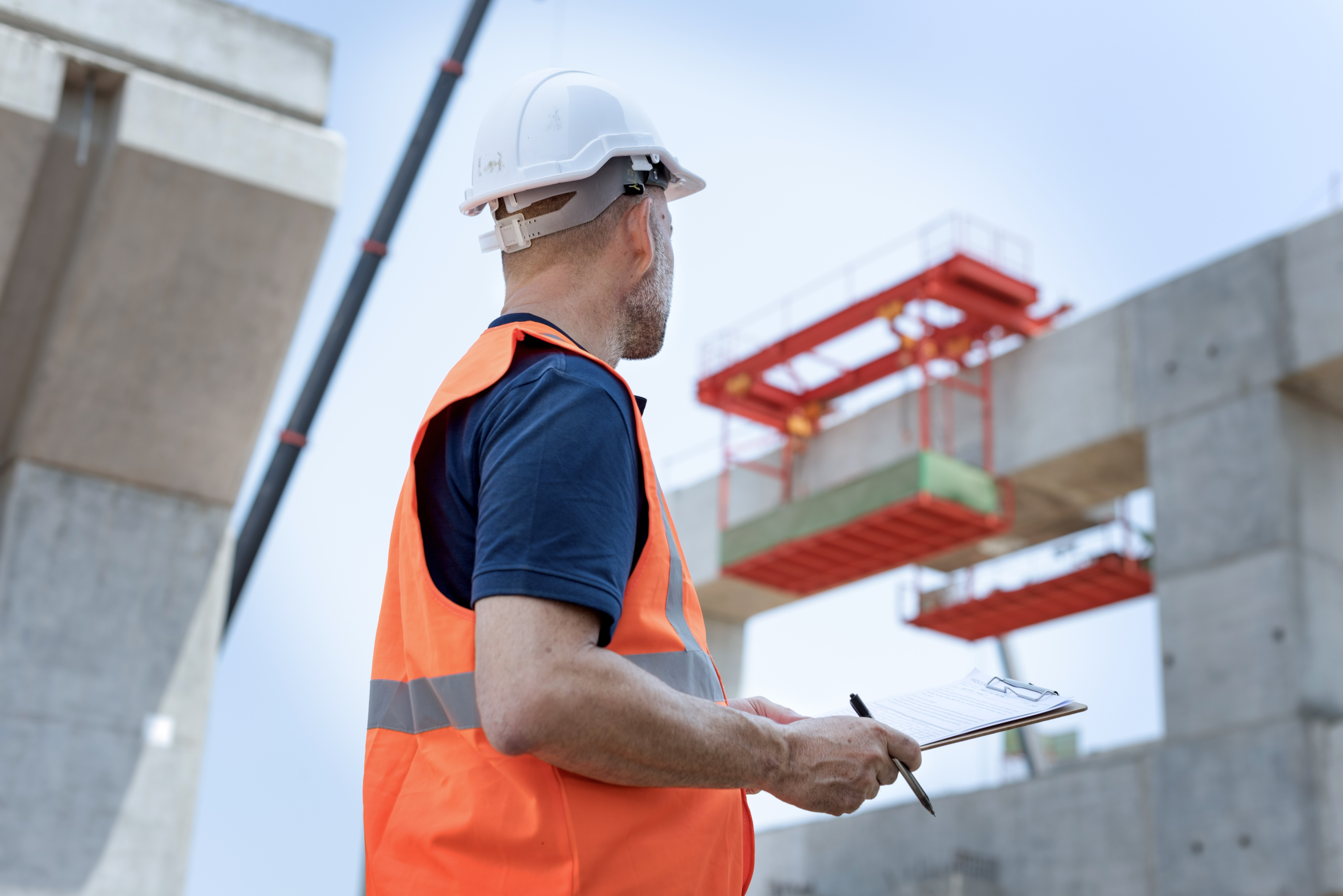 A man is looking at a construction site, wearing a reflective vest and safety helmet, holding a clipboard and a pen.